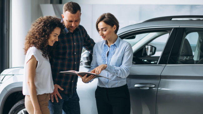 Três pessoas em uma concessionária observam um catálogo ao lado de um SUV prateado; uma delas, possivelmente a vendedora, aponta informações no material enquanto o casal escuta atentamente.