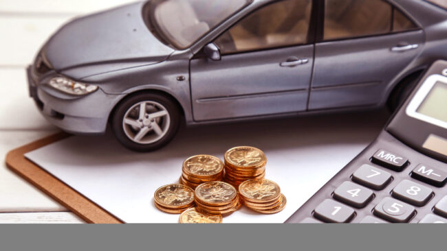 Imagem com miniatura de carro cinza, pilha de moedas douradas, calculadora e prancheta sobre superfície branca, representando planejamento financeiro automotivo.